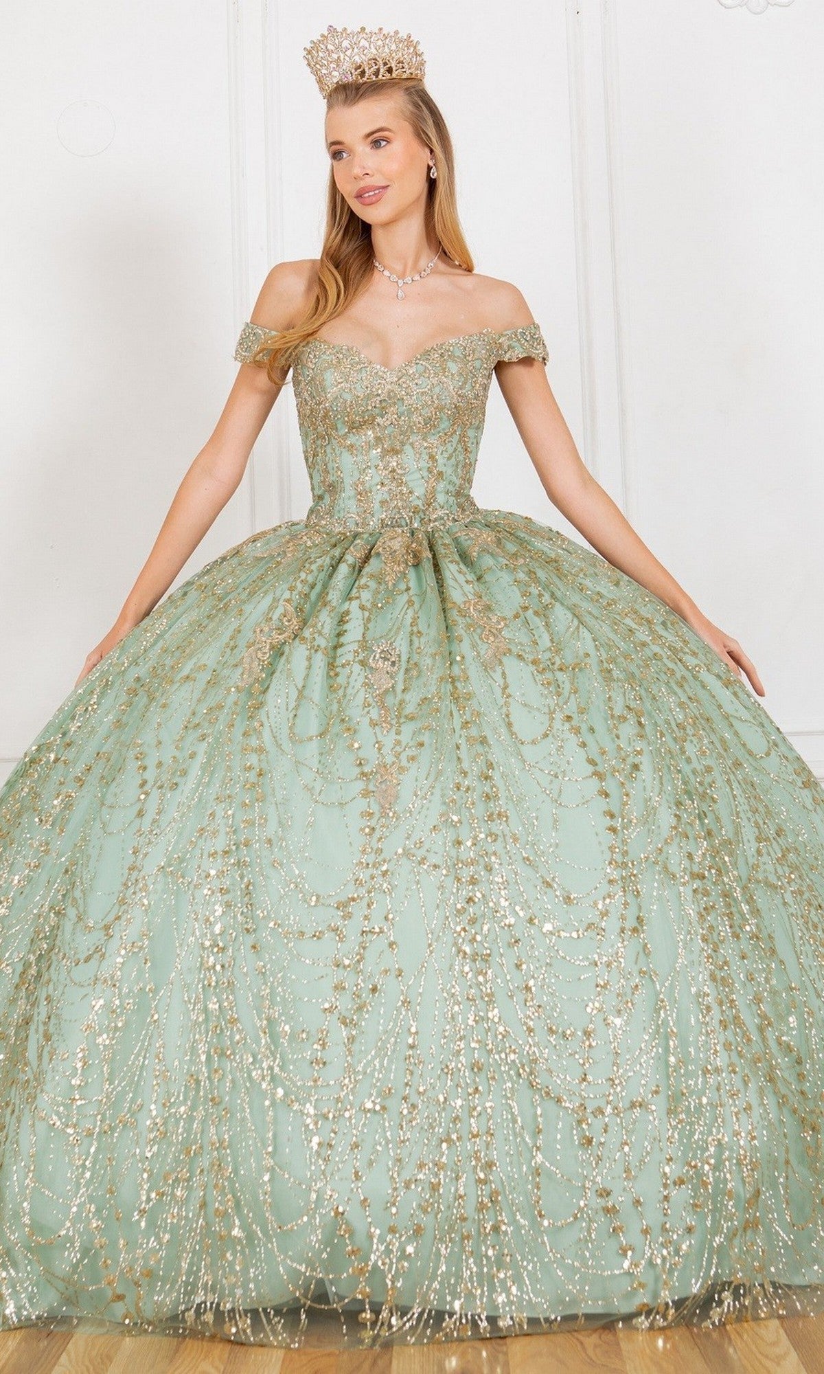 A woman wearing the Quinceanera Dress 4201 By Cinderella Couture in sage, an off-the-shoulder ball gown adorned with gold embellishments and paired with a crown, stands in front of a white wall.